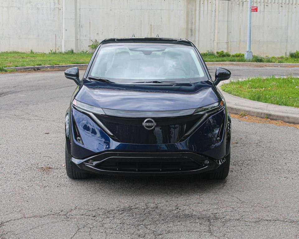 A blue Nissan Ariya electric SUV seen from the front, against a concrete wall.