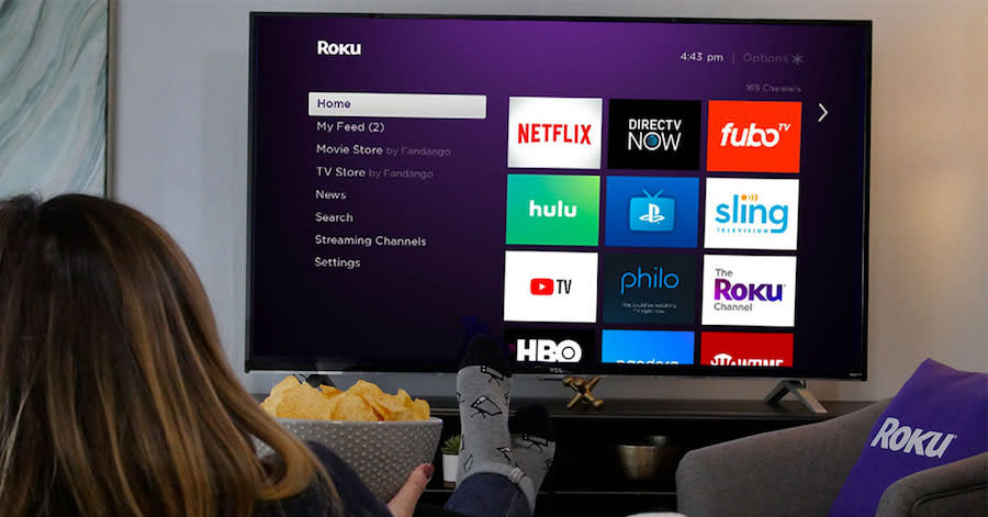 A television displaying the Roku menu and a woman holding a bowl of chips watching from the sofa