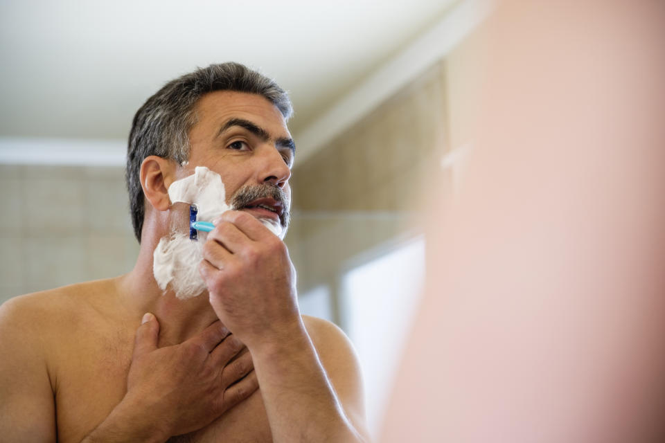 A shirtless man uses a razor to shave his face, looking in the mirror with focused attention. The bathroom setting is visible in the background