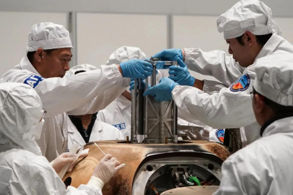 Laboratory technicians in clean white suits remove a metal container from a charred space capsule