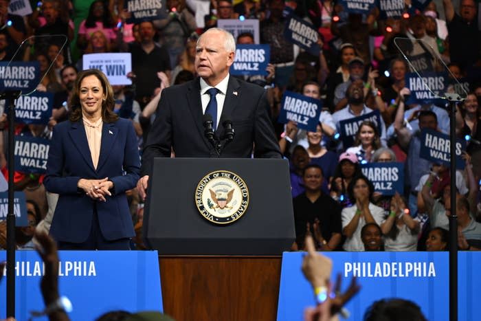 Kamala Harris in a suit and Tim Walz at a podium with a seal, speaking at a rally in Philadelphia. Supporters hold signs that read "Harris Walz" and "Together We Win"