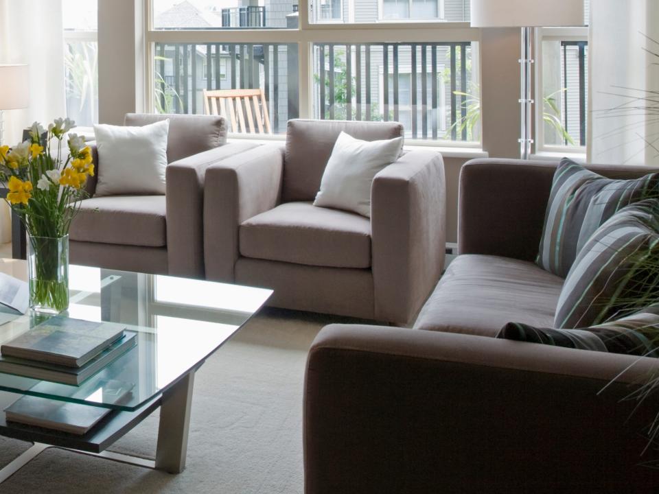 Living room with brown two brown sofas and window with view of cityscape surrounding a glass table with a vase on top in front of a white fireplace and large flatscreen tv