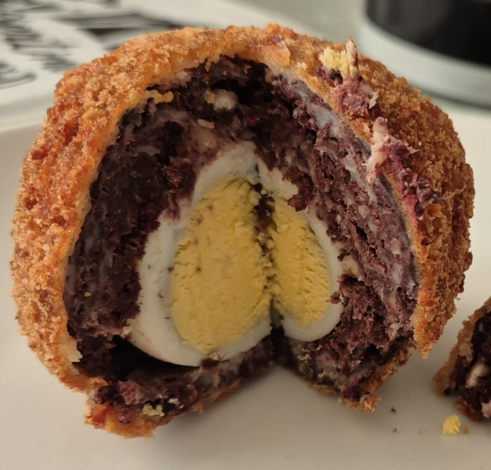 A halved Scotch egg is displayed on a plate, revealing its breaded outer layer, sausage meat, and a hard-boiled egg at the center