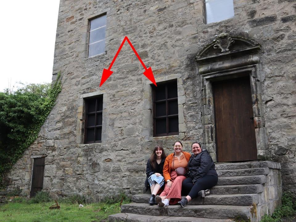 A group of three people sitting on the exterior stairs of Midhope Castle in Scotland, with two arrows pointing at a pair of modern windows installed in the historical building.