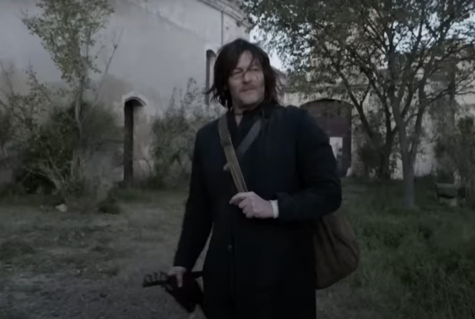 Norman Reedus as Daryl Dixon in The Walking Dead: Daryl Dixon stands outside a building, holding a crossbow and wearing a coat with a bag over his shoulder