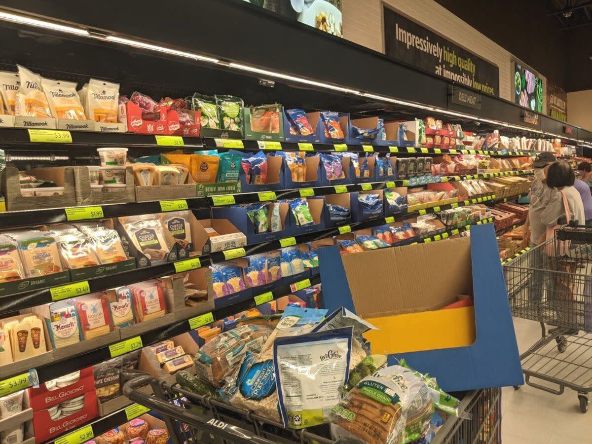 Cheese refrigerated section at Aldi, Chandler, Arizona, six rows of many different kinds of cheeses with an overflowing shopping cart in the foreground, lunch meats section in the background