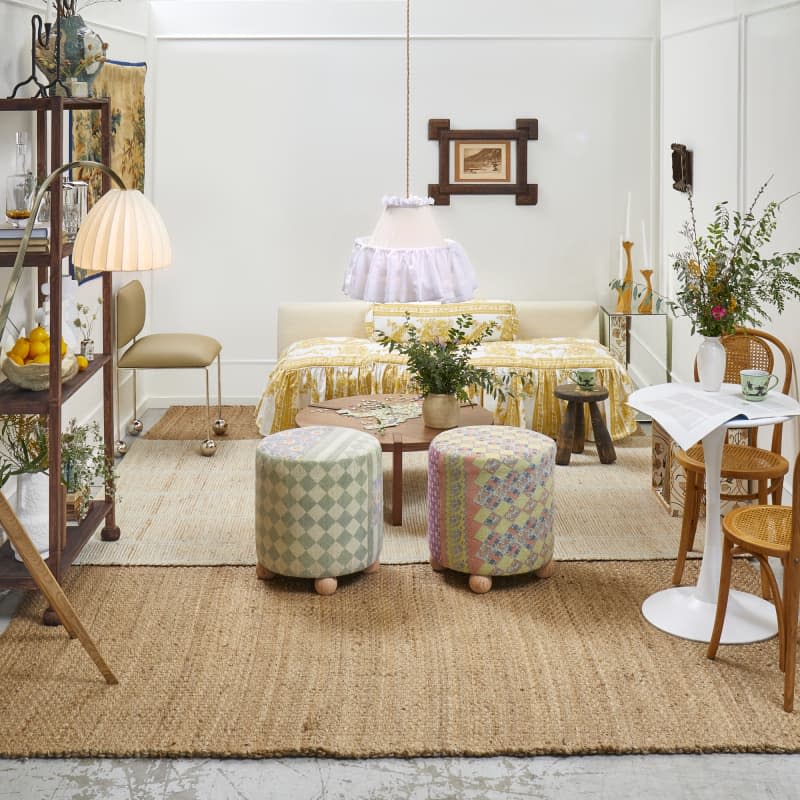 Head on view of a living room with white walls, a low couch with printed yellow and white ruffled cushions, two patchwork stools, and a small bistro table and chairs in the front right.