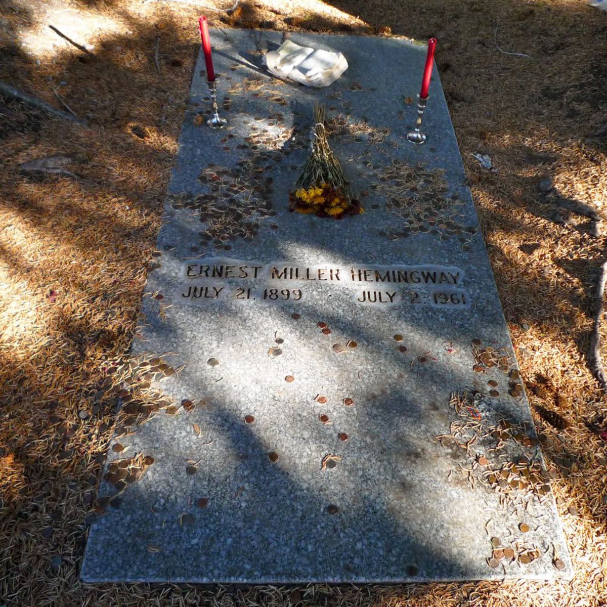 Ernest Hemingway's headstone in the Ketchum Cemetery, Ketchum, Idaho with coins on it, two candle sticks and a few dried flowers, surrounded by fallen brush with the sunlight scattered