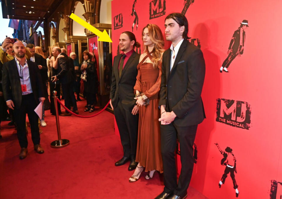 Three individuals at an event, the middle person in a rust-colored dress with shoulder cut-outs, flanked by two people in suits