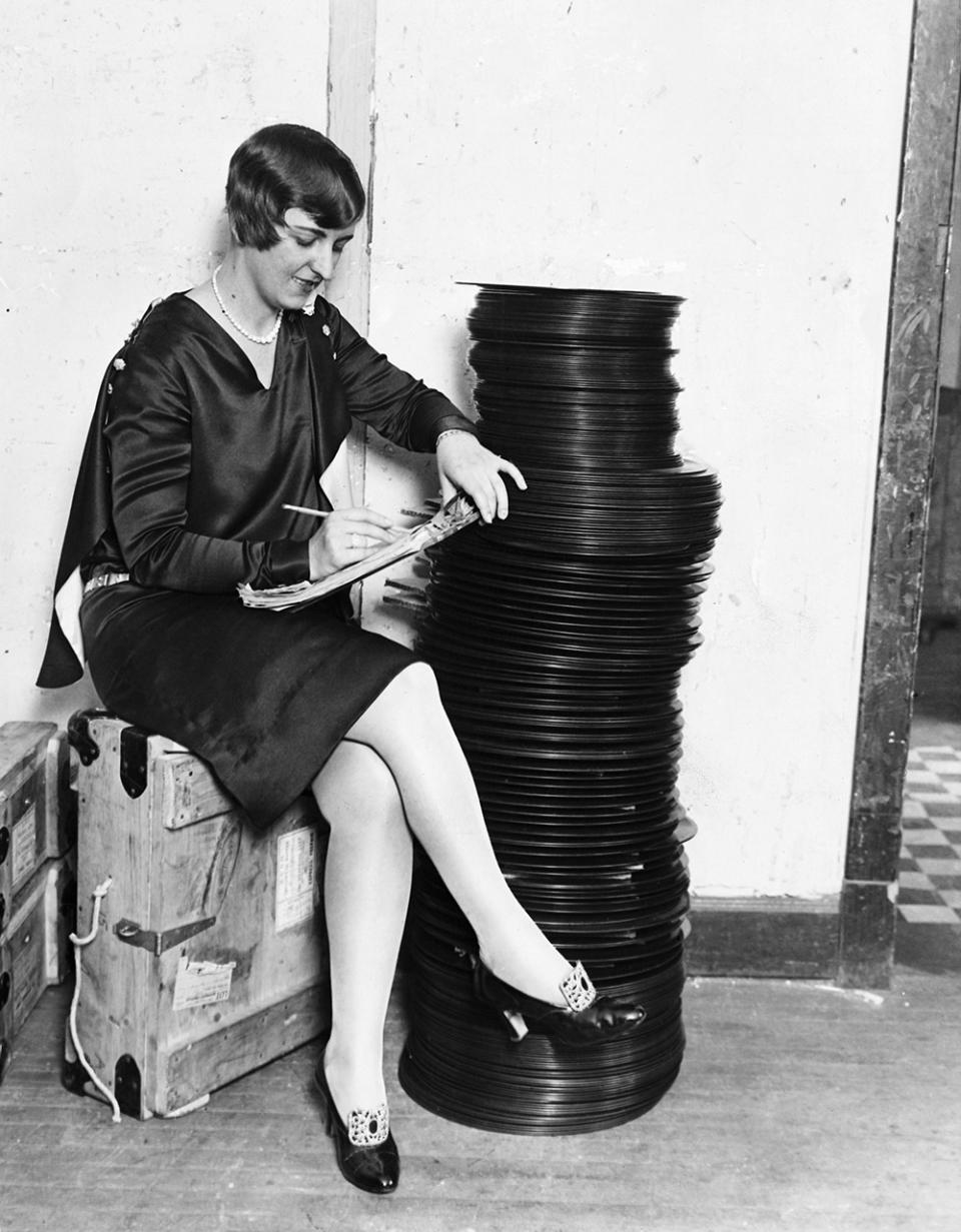 Our photo shows Miss Tess Horaty checking up on Vitaphone Records at the company's place in Chicago. Since the installation of the machines in various theaters it is extremely hard for the Vitaphone company to meet the supply and demand for the records.