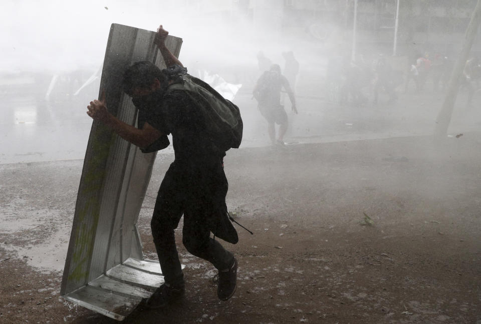 An anti-government protester uses a piece of sheet metal to protect himself from the jet spray of a police water cannon in Santiago, Chile, Friday, Nov. 8, 2019. Chile's president on Thursday announced measures to increase security and toughen sanctions for vandalism following three weeks of protests that have left at least 20 dead. (AP Photo/Esteban Felix)