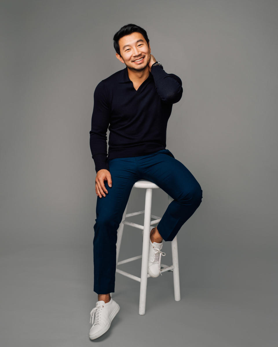 Simu sitting on a stool with his hand on his neck as he smiles
