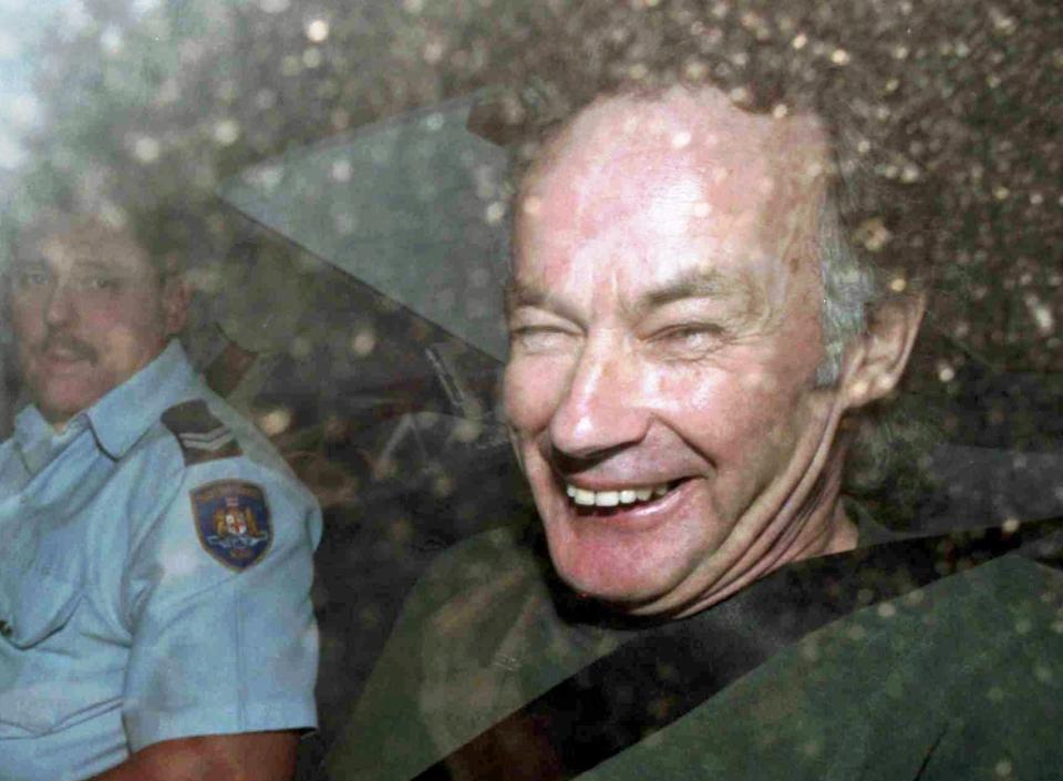 Picture of serial killer, Ivan Milat, smiling while sitting with a police officer in the backseat of a car. 
