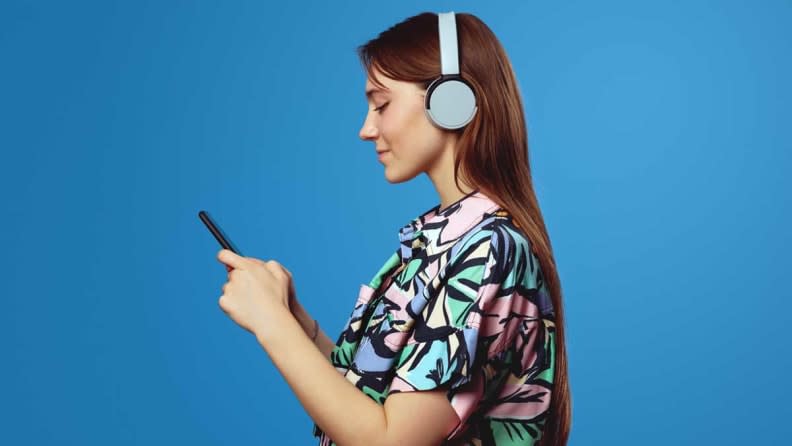 Woman wearing headphones listening to content on her smartphone in front of a blue background.