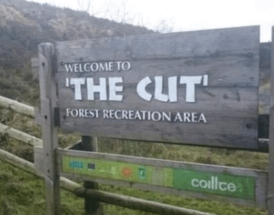 A wooden sign reading "Welcome to 'The Cut' Forest Recreation Area," but the u in "The cut" makes it look like it says "Welcome to 'The Clit'"