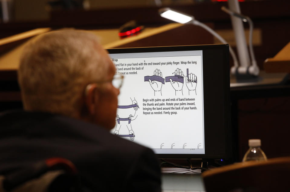 Former U.S. Sen. Harry Reid sits in front of a monitor showing instructions for an exercise band in court Tuesday, March 26, 2019, in Las Vegas. A jury in Nevada heard opening arguments Tuesday in Reid's lawsuit against the maker of a flexible exercise band that he says slipped from his hand while he used it in January 2015, causing him to fall and suffer lasting injuries including blindness in one eye. (AP Photo/John Locher)