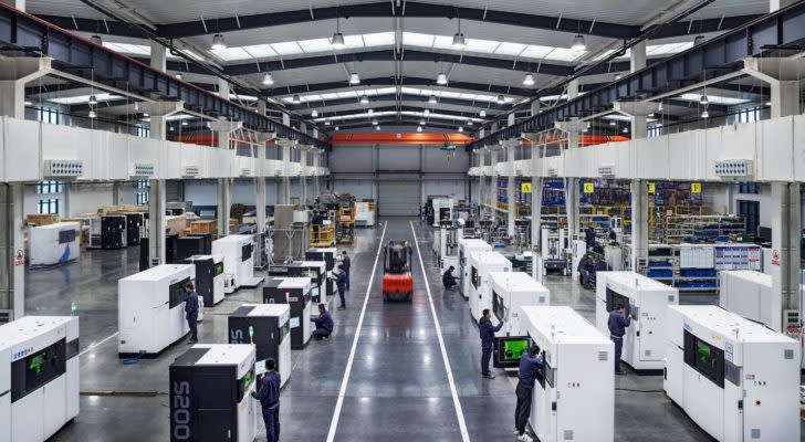 A picture of multiple 3D Printers with people working on them in a factory.