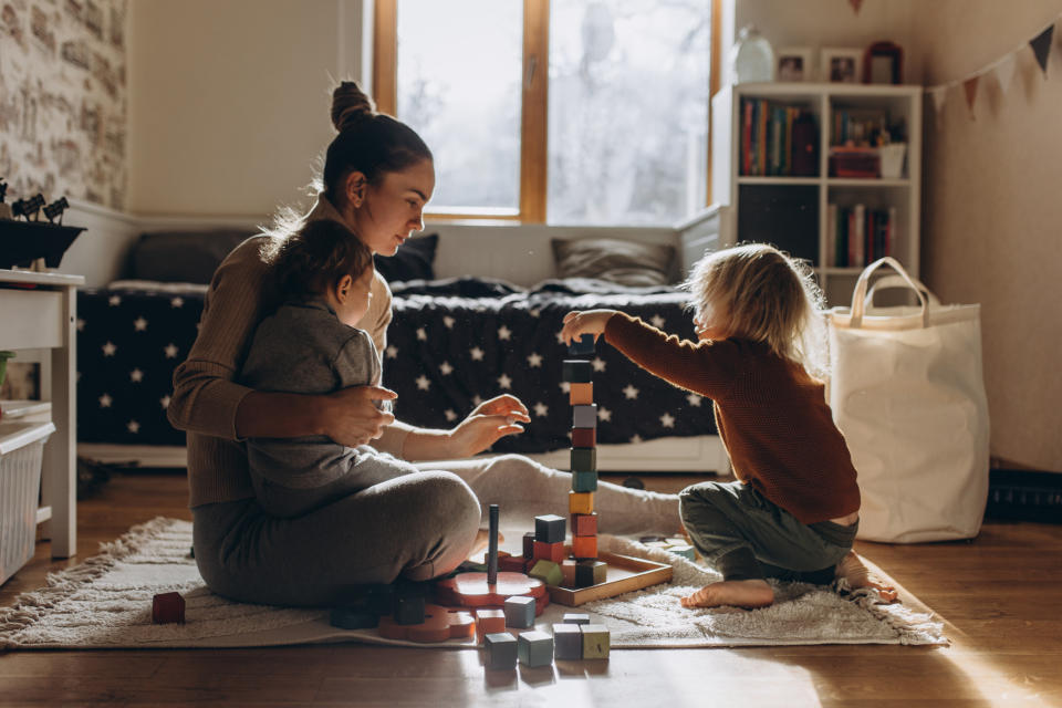 A parent sits on the floor with two children, one in their arms, while the other child stacks colorful blocks in a cozy, sunlit room