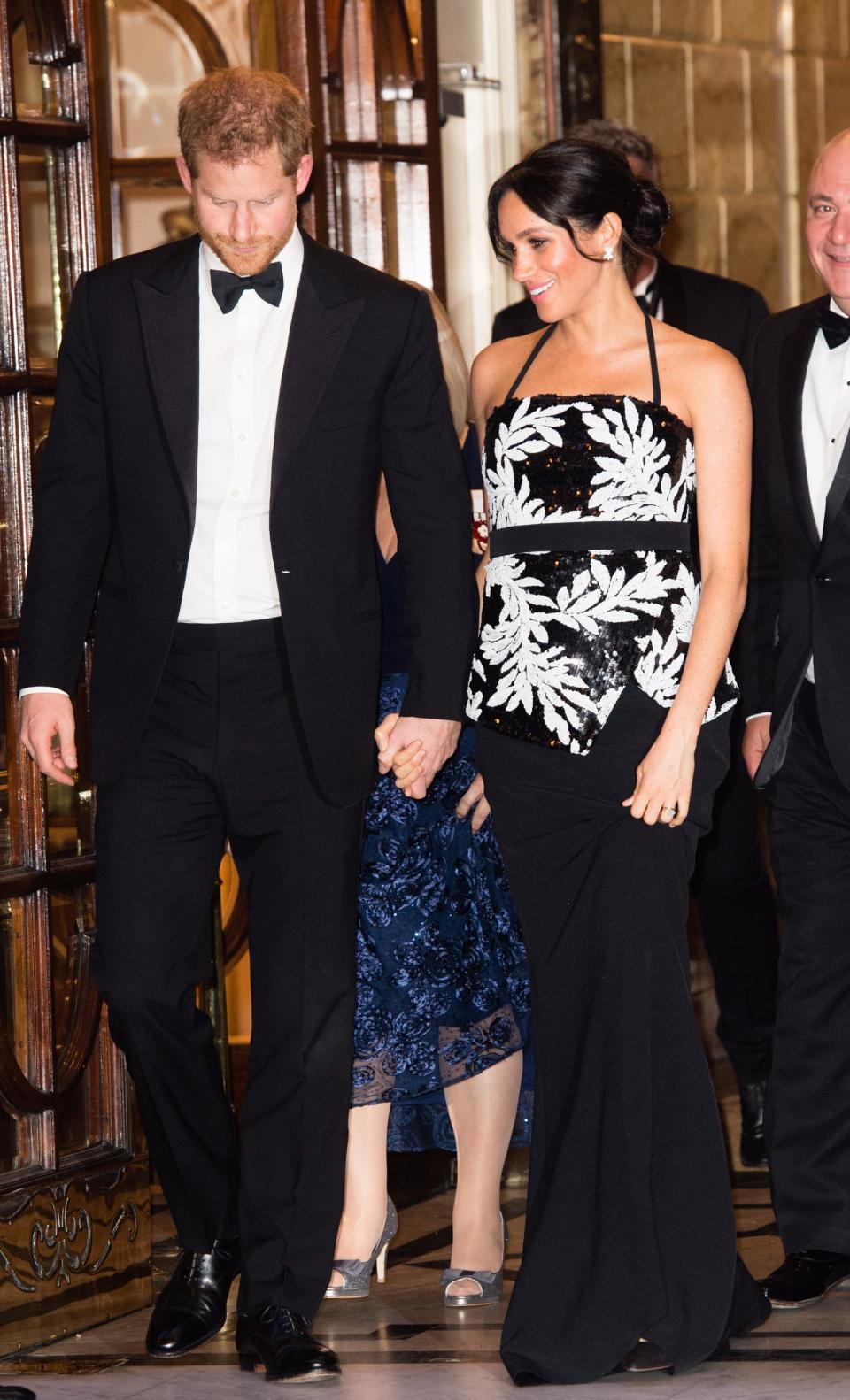 Harry in a black tuxedo and bowtie and Meghan in a white palm leave print black top and black floor length skirt.