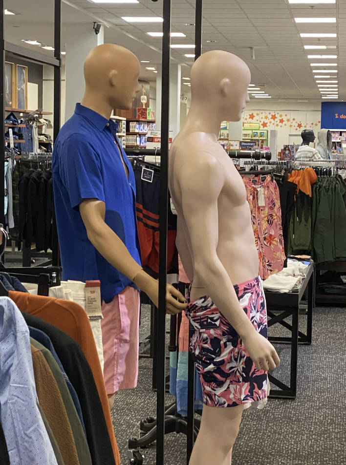 Two mannequins in a store, one dressed in a blue shirt and pink shorts, the other shirtless wearing floral shorts. Shelves with clothing in the background