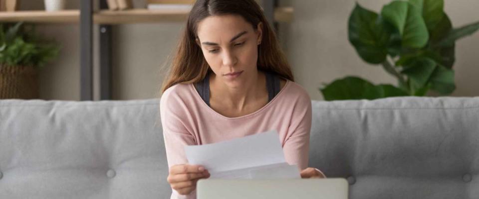 Upset woman sitting on couch at home near computer holding paper reading bad news about her student loan debt.