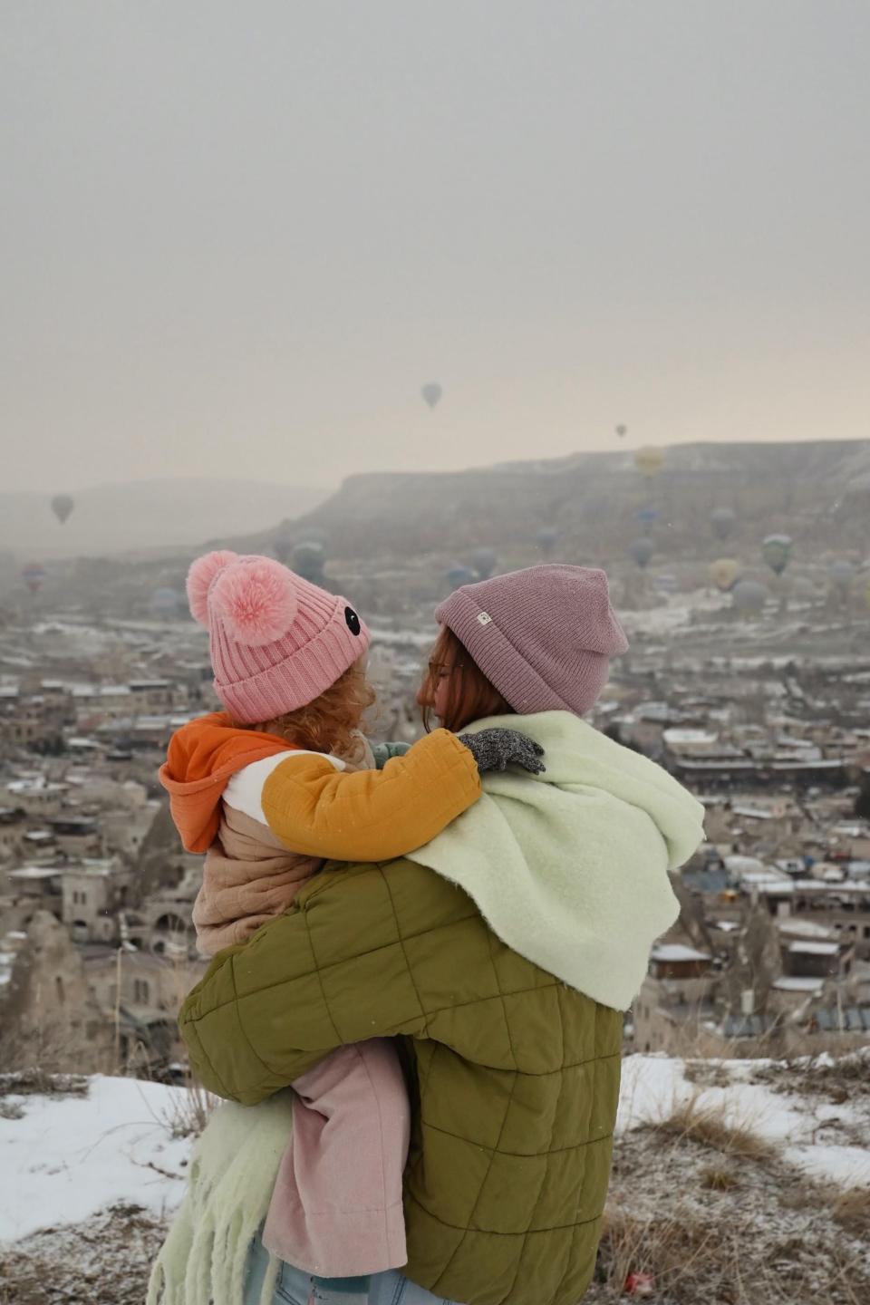 A daughter looks at her mother in front of a city with hot air balloons floating over it.