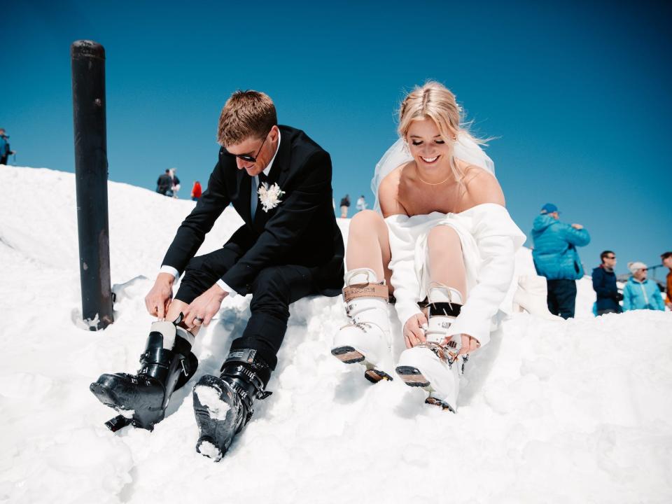A bride and groom put on ski boots on a snowy mountain.