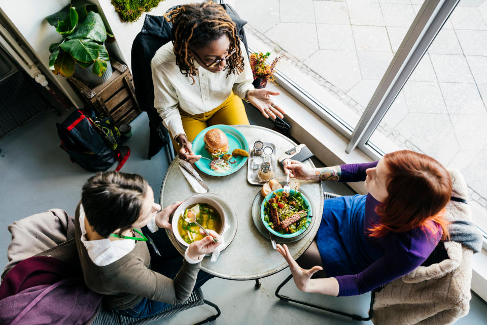 An aerial view of three women sitting at a small table in a vegan cafe, eating colorful plates of food.