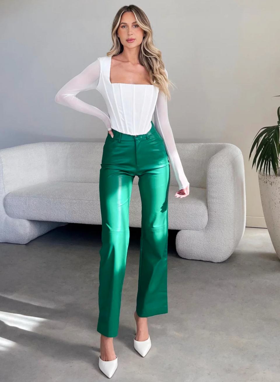 A  model with long blonde hair worn loose wears bright green faux leather pants over a white top with square neckline and long sleeves. She stands in a loungeroom setting with white lounge and large potted palm.