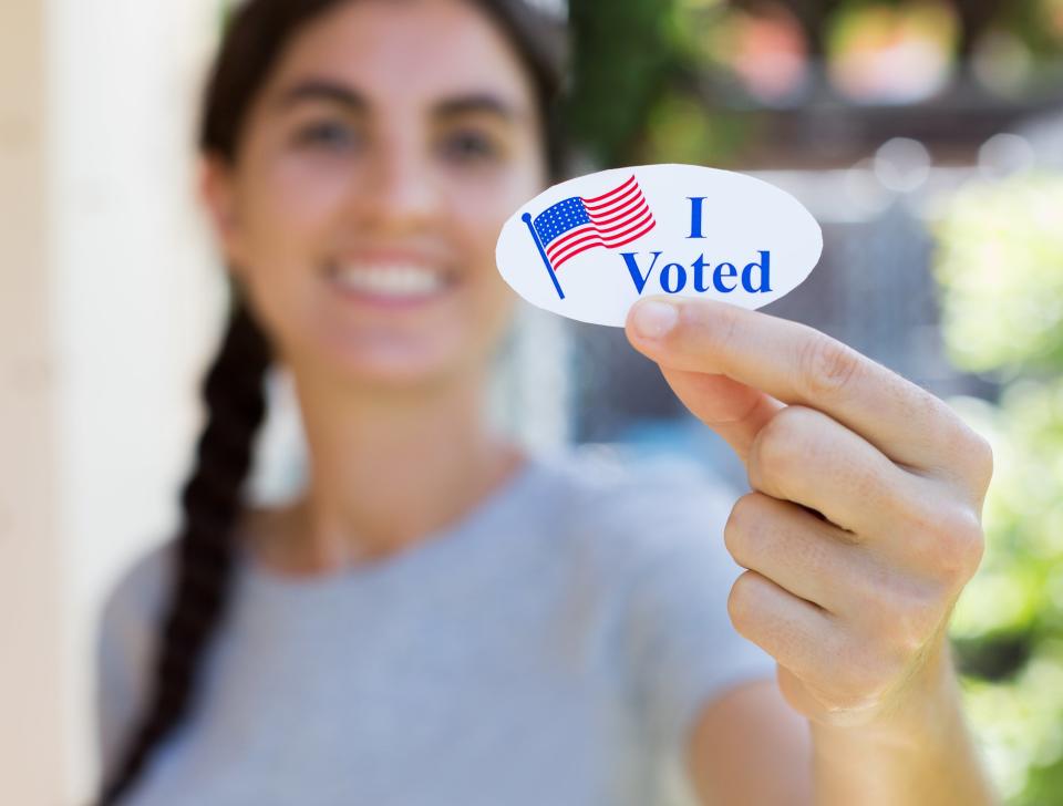 A woman holding up an "I Voted" sticker, smiling