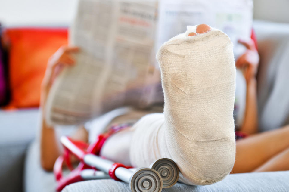 Person with a bandaged leg reading a newspaper while lying on a couch with crutches nearby