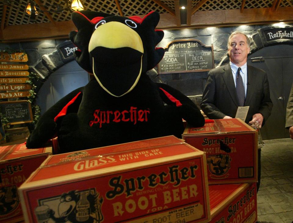 Rooty the Griffin, the Sprecher Root Beer mascot, stands surrounded by boxes of root beer as Howard Dean, then a Democratic presidential candidate, stands in the background in 2004.