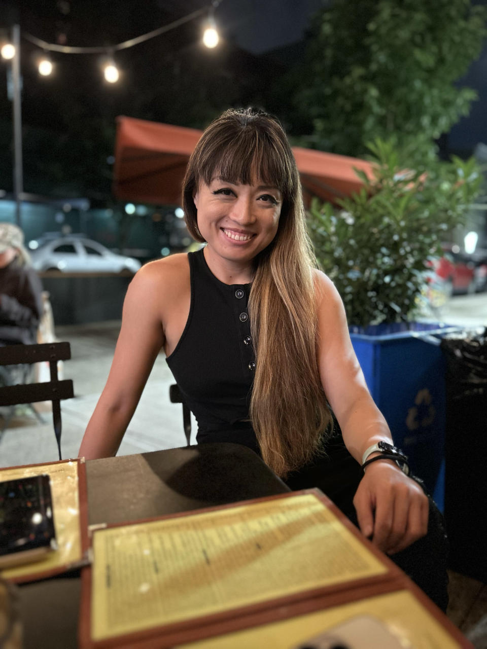 <p>A photo from the iPhone 14 showing a woman sitting at a table at an outdoor restaurant, smiling. The background around her is slightly blurred.</p>
