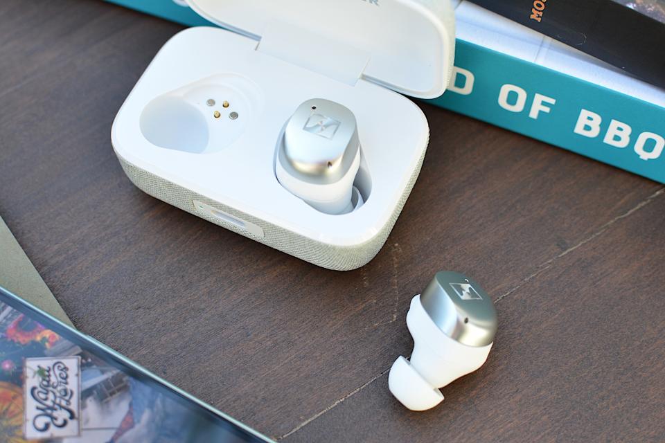 <p>White/silver Sennheiser Momentum True Wireless 4 earbud sitting in its case with the lid open, one earbud sitting on a wooden table on its side.</p>
