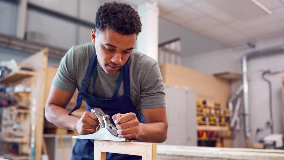 Male Student Studying For Carpentry Apprenticeship At College Using Wood Plane.