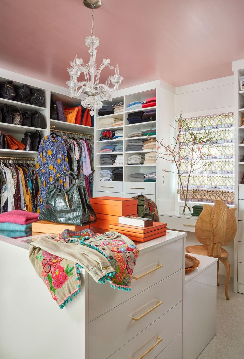 walk in closet with a leaf back chair next to a window and lots of white drawers and shelving for clothes and a colorful jacket with blue pompoms draped over the drawers in the foreground as well as a archive boxes and other clothes
