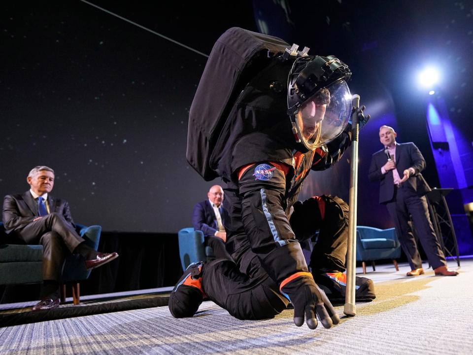 man in black spacesuit kneels to touch the floor on a stage