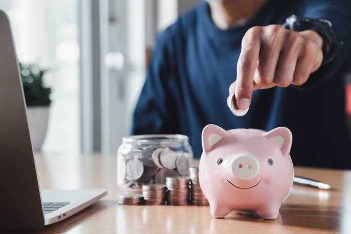 Person saving money by placing a coin into a piggy bank, with a jar of coins and laptop on the desk