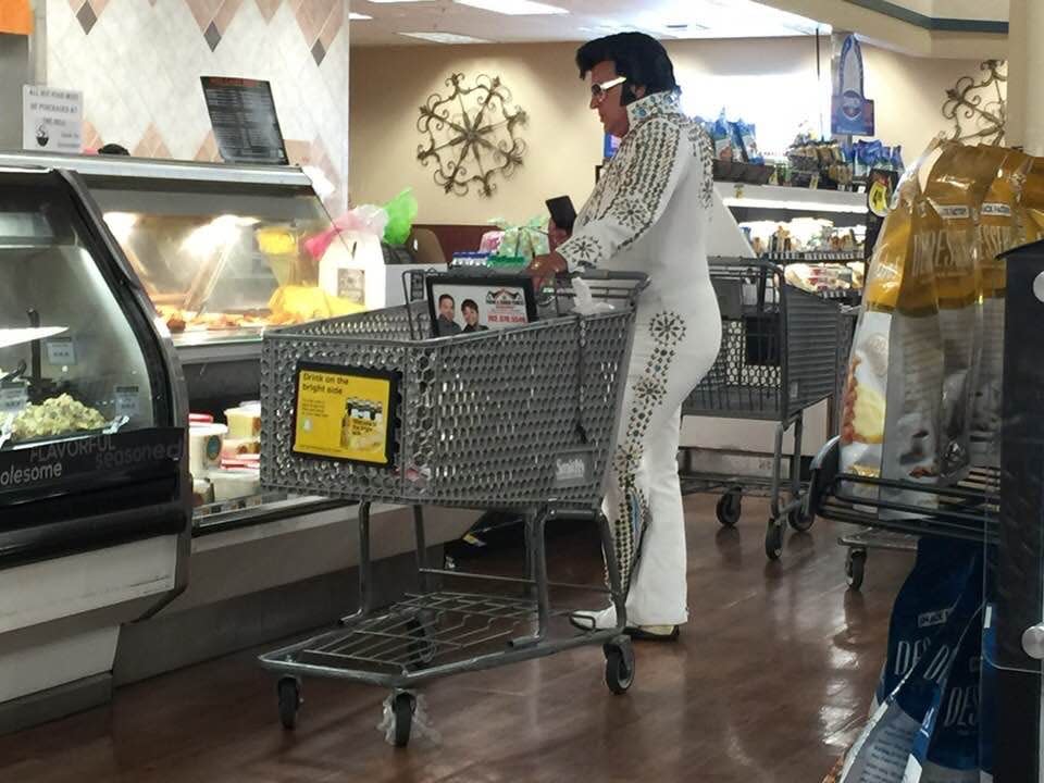 An Elvis Presley impersonator pushing a shopping cart while looking at a shelf of produce in a grocery store.