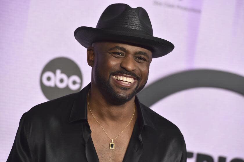 Wayne Brady smiles while wearing a black fedora and a black button up that reveals his chest and a gold necklace