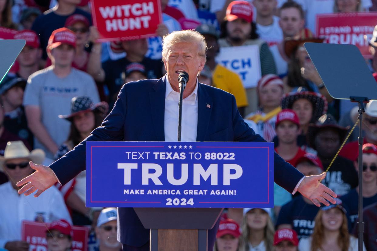 Former President Donald Trump surrounded by supporters on bleachers holding signs saying: Witch Hunt. A sign on the podium in front of him says: Trump, Make America Great Again! 2024.