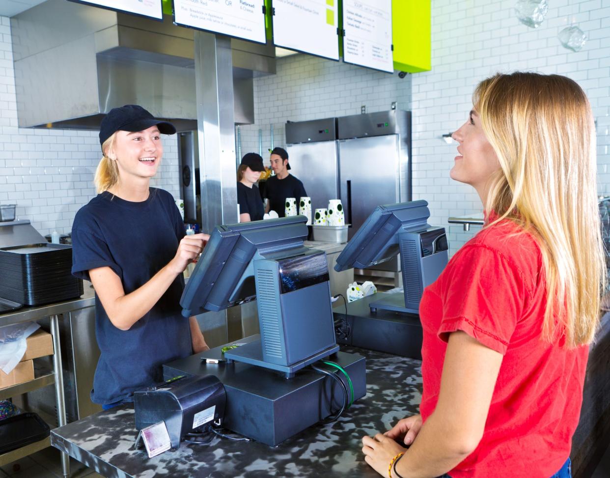 A young woman customer placing her order at a fast food convenience restaurant. A young woman server staff is assisting her at the checkout cashier counter with the kitchen staff working in the background.