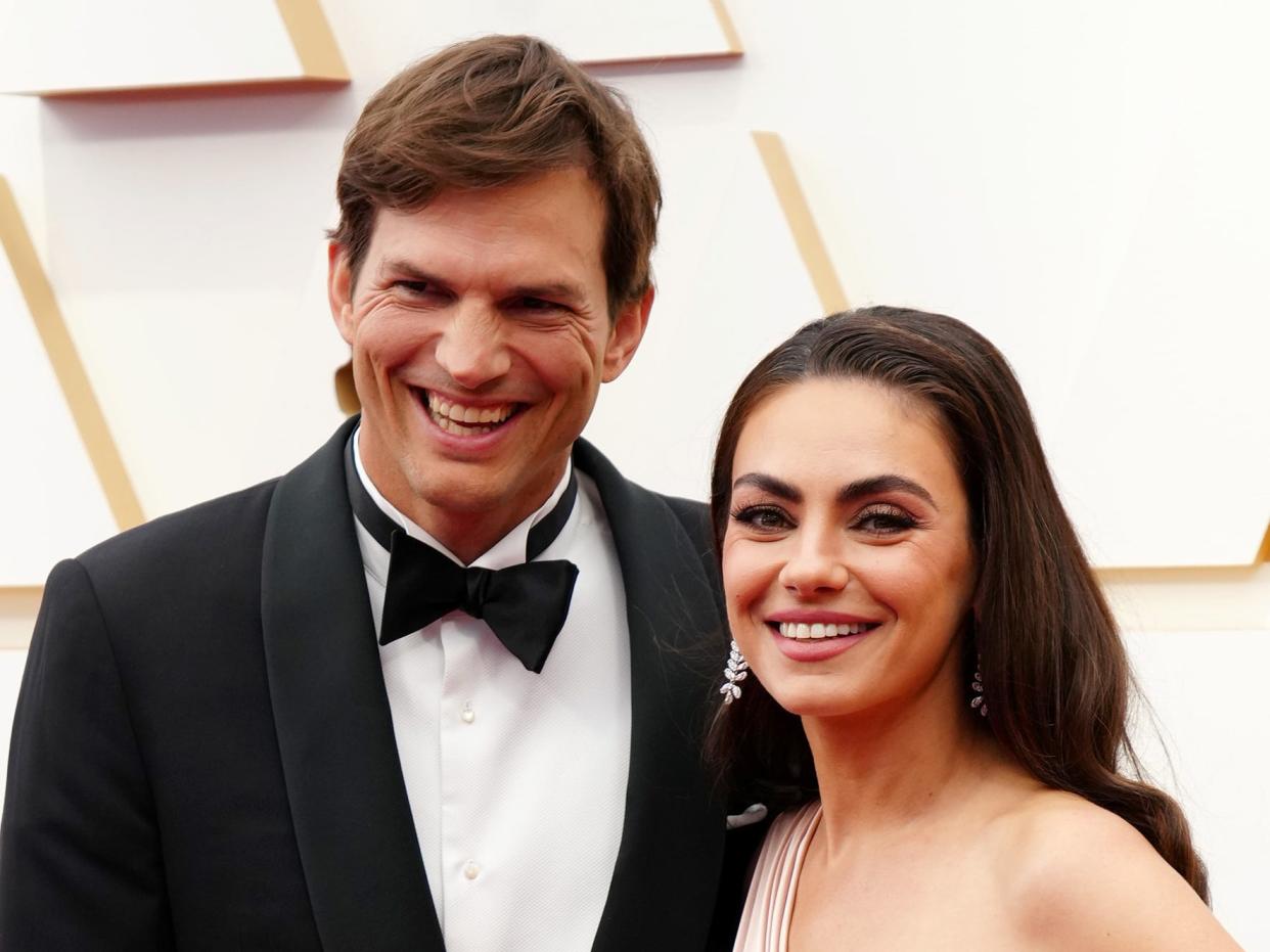 Ashton Kutcher, in a tuxedo, and Mila Kunis, in a strapless blush gown, smile while posing on the red carpet together.