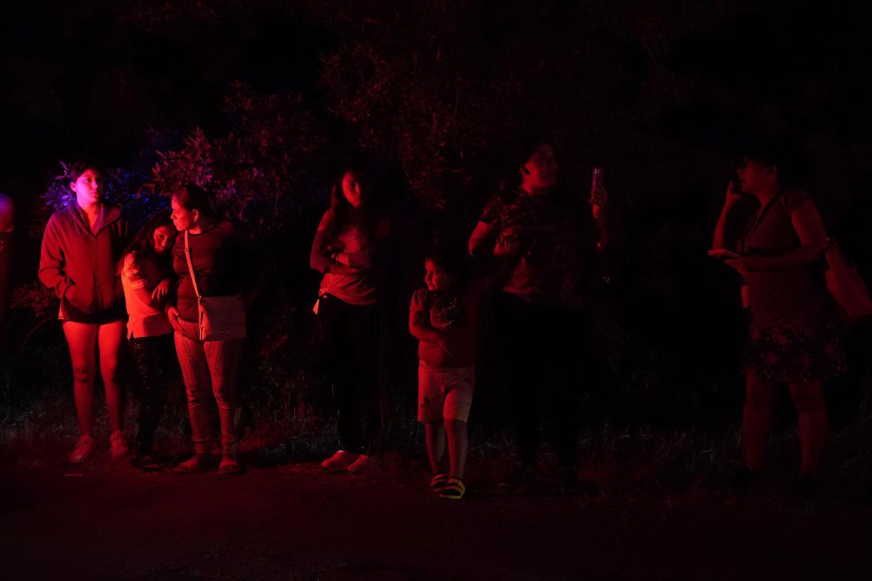 Onlookers are illuminated by police lights at the scene where officials say dozens of people have been found dead and multiple others were taken to hospitals with heat-related illnesses after a semitrailer containing suspected migrants was found on Monday, June 27, 2022, in San Antonio.