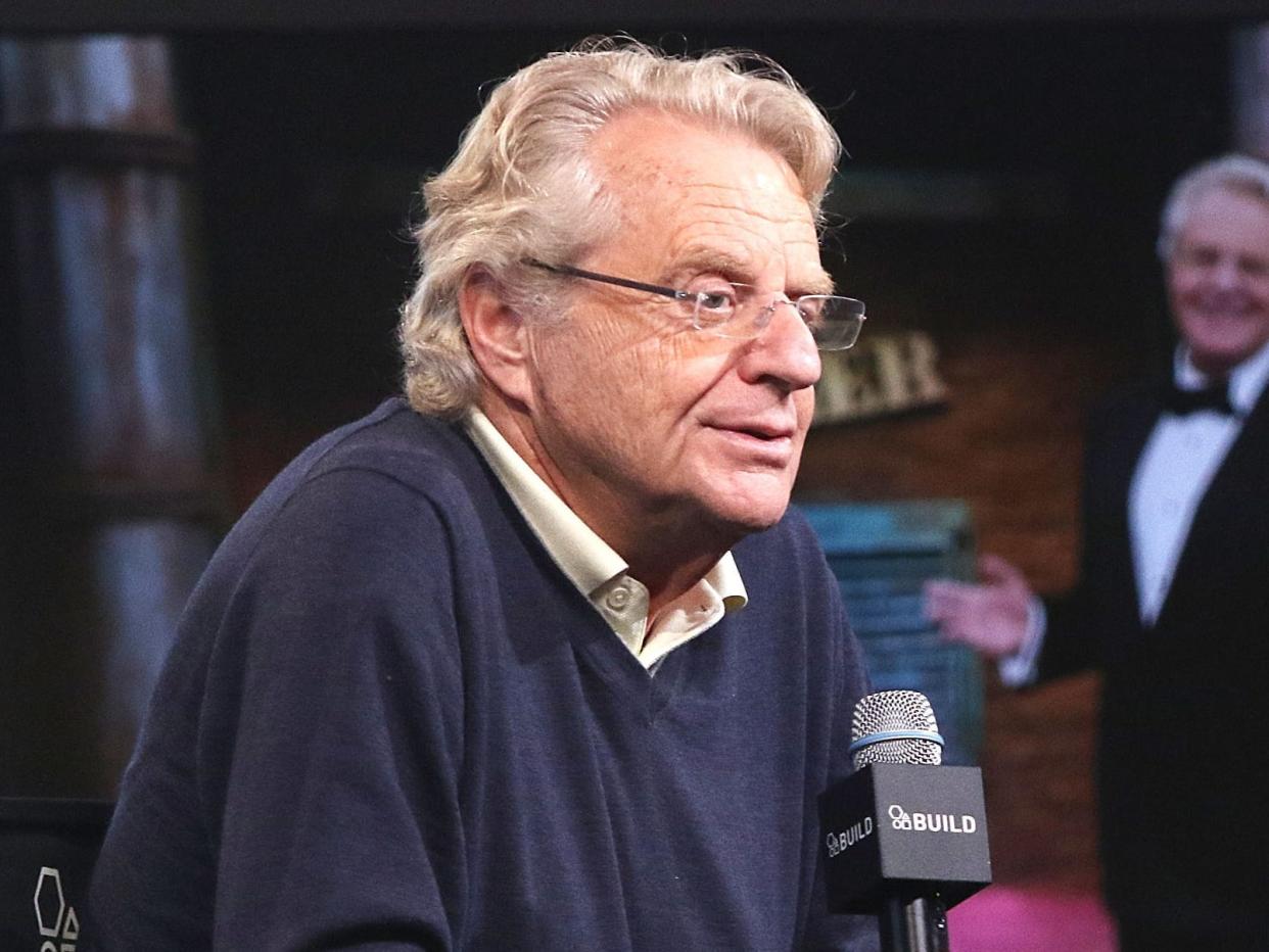 Jerry Springer speaking into a microphone while a man in a black bowtie and suit gestures towards him.