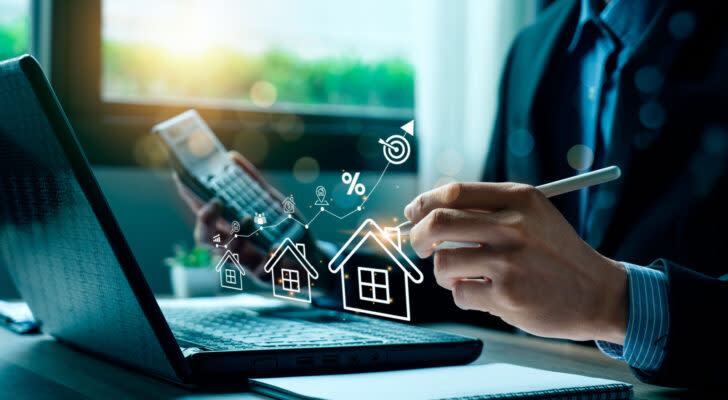 Business person at laptop computer holding a calculator and pen while graphic of houses and upward chart floats above computer