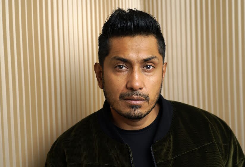 Tenoch Huerta poses in a dark green velvet jacket and black shirt against a gold background.