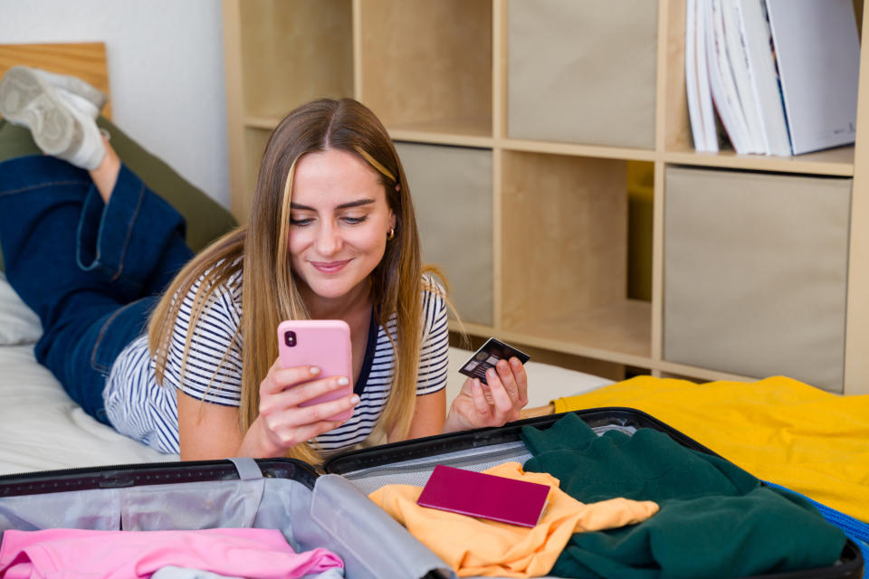 A woman lies on a bed, smiling while using her phone and holding a credit card. An open suitcase with clothes and a passport is in front of her