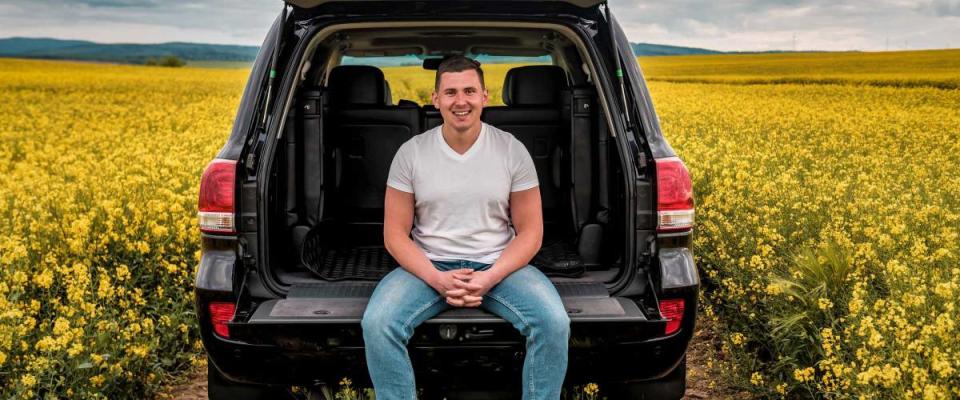 smiled young man sitting in the trunk of a car in yellow rape field