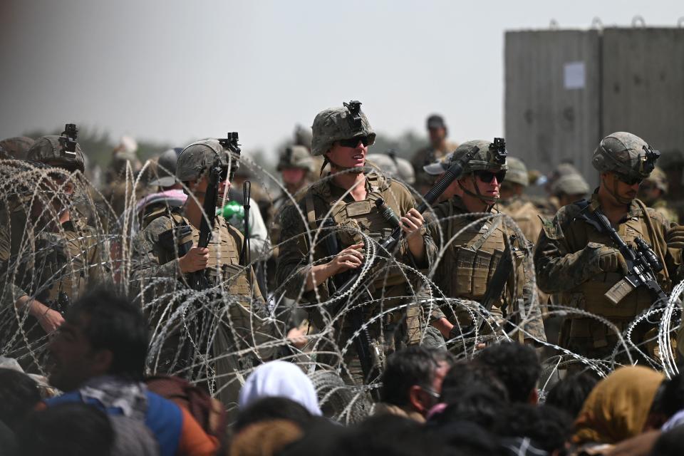 U.S. soldiers stand guard behind barbed wire as Afghans sit on a roadside near the military part of the airport in Kabul on August 20, 2021. / Credit: WAKIL KOHSAR/AFP via Getty Images